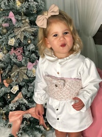 a little girl in a white dress standing in front of a christmas tree