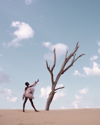 a girl in a dress is standing in the sand next to a dead tree